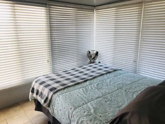 A bed with a green quilt and checkered blanket in a room with white blinds on the windows.