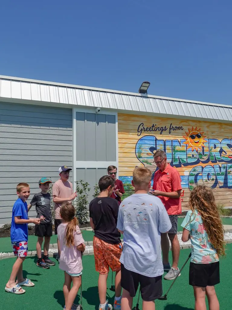 A group of children and an adult are gathered around a mini golf putting green in front of a building with a mural that reads 'Greetings from Sunders' with a sun logo.