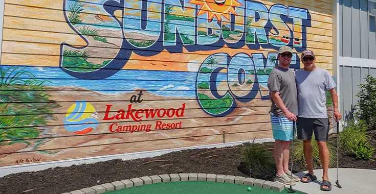Two men standing in front of a mural with the words 'Sunburst Cove at Lakewood Camping Resort'