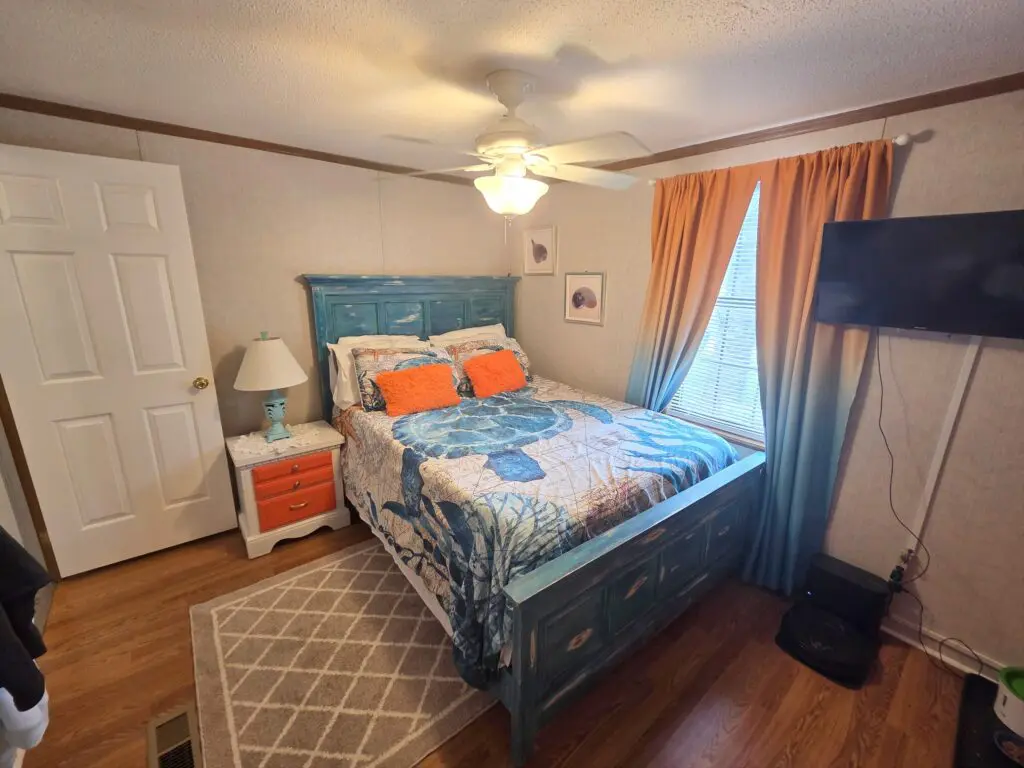 A wide-angle view of a bedroom with a blue bed, white and orange nightstand, and a large TV on the wall.
