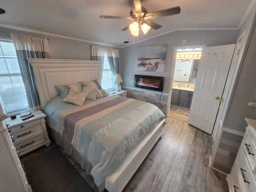 A wide-angle view of a hotel room with a large bed, a ceiling fan, a fireplace, and a doorway leading to another room.